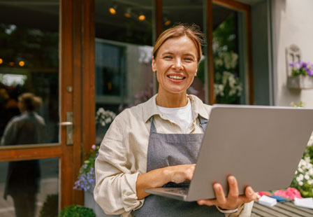 Smiling woman in an apron standing with a laptop outside a business